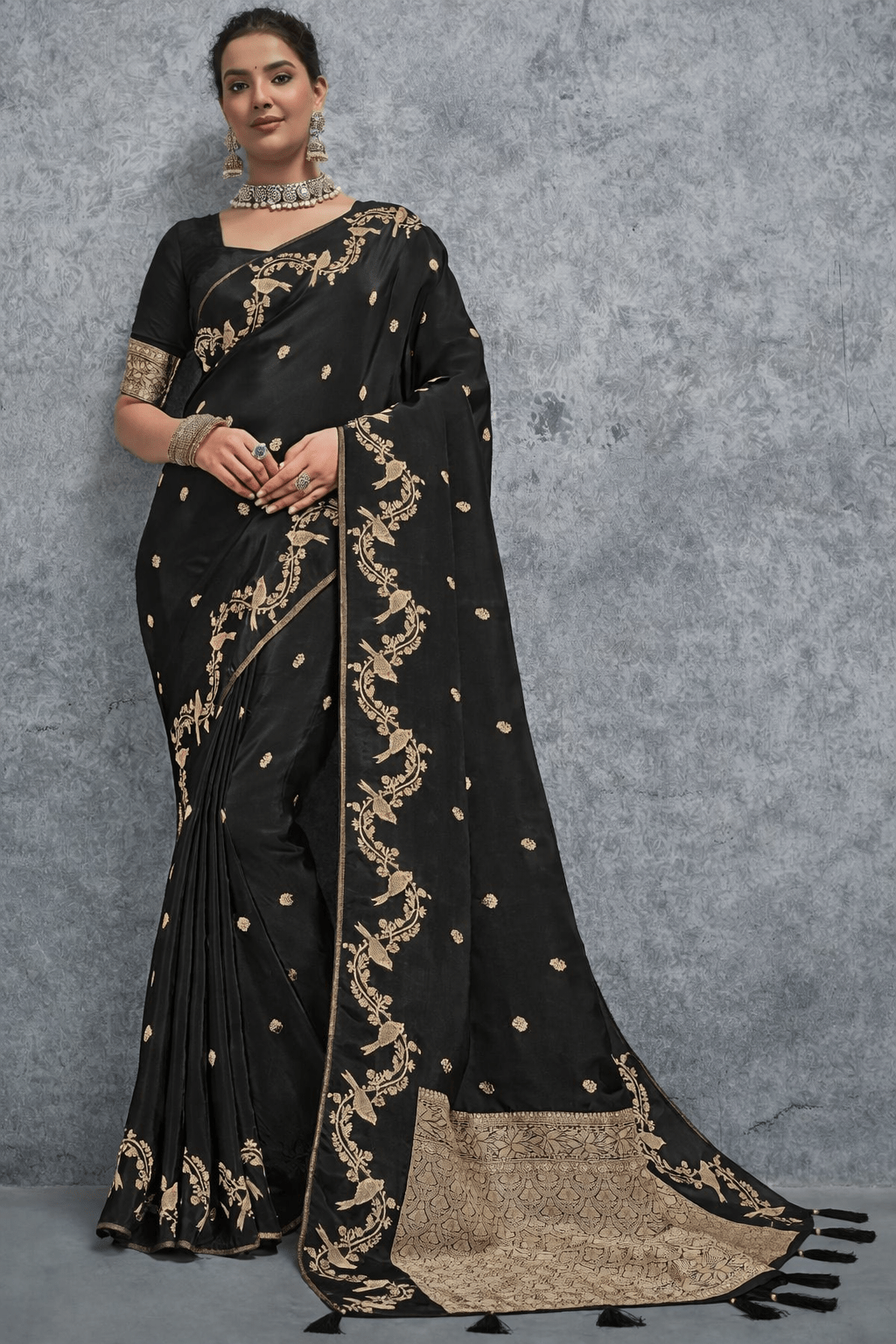 Black silk saree with intricate gold zari embroidery, featuring floral vine motifs and a richly woven traditional border, elegantly draped on a model against a grey textured background – perfect for wedding and festive wear.