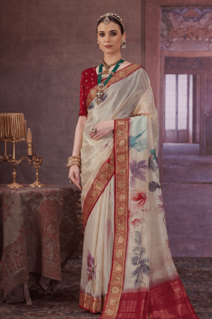Woman wearing an off-white silk saree with rich red and gold zari border and pastel floral prints, paired with a red blouse and traditional jewellery in a royal indoor setting.